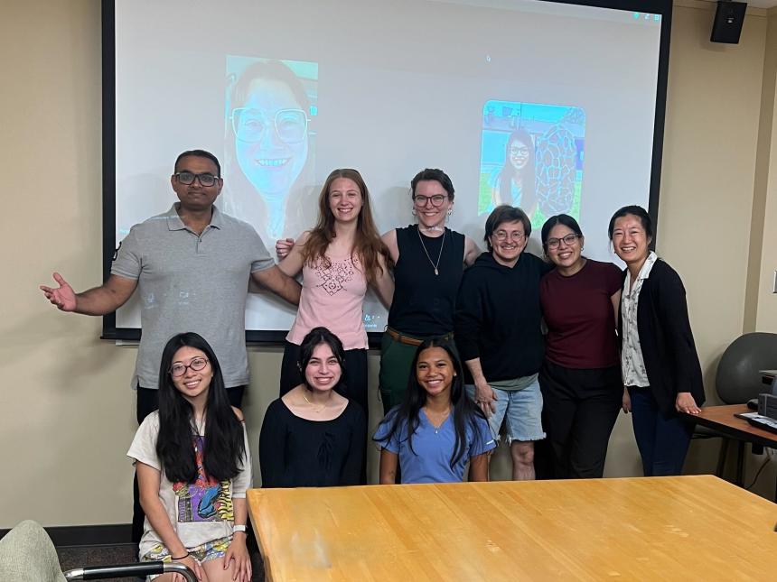 A group of 8 people in a conference room in front of a projector screen that has the face of Nicole